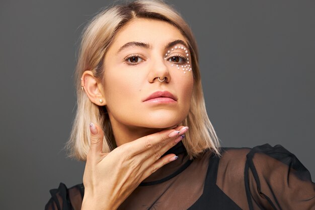 Mujer rubia muy de moda con maquillaje glamoroso de moda posando aislado sosteniendo la mano debajo de la barbilla, mostrando uñas pulidas y pulidas con expresión facial enigmática segura