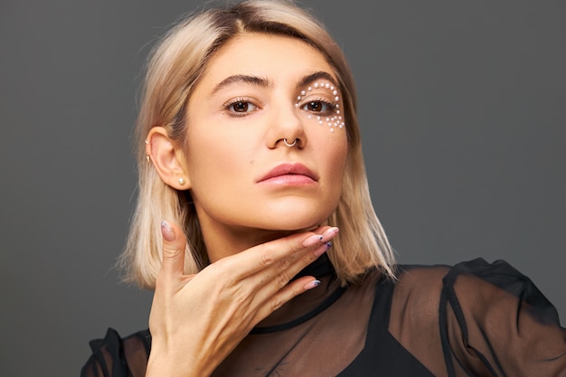 Mujer rubia muy de moda con maquillaje glamoroso de moda posando aislado sosteniendo la mano debajo de la barbilla, mostrando uñas pulidas y pulidas con expresión facial enigmática segura