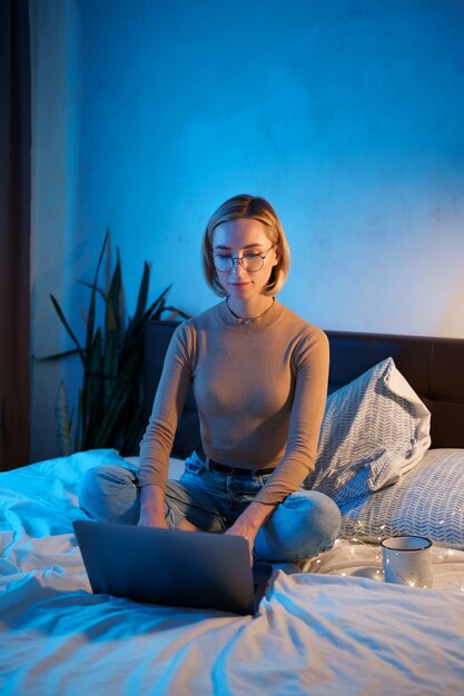Mujer rubia de pelo corto en la cama blanca en jeans con una computadora portátil.