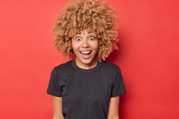 Mujer rubia de pelo rizado mira con feliz expresión sorprendida sonríe ampliamente vestida con una camiseta negra casual aislada sobre un fondo rojo brillante reacciona ante una oferta inesperada Reacciones humanas