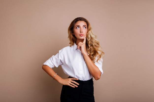 Mujer rubia pensativa que presenta el aislante en la pared beige. Ropa de trabajo casual con estilo.