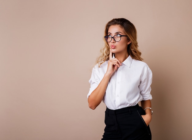 Mujer rubia pensativa que presenta el aislante en la pared beige. Ropa de trabajo casual con estilo.