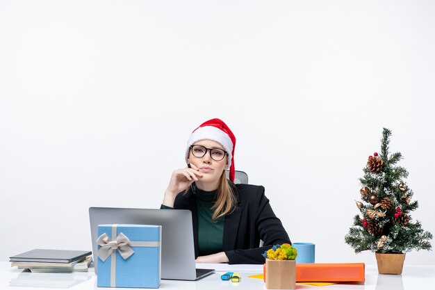 Mujer rubia pensativa con un sombrero de santa claus sentado en una mesa con un árbol de Navidad y un regalo en la oficina sobre fondo blanco.