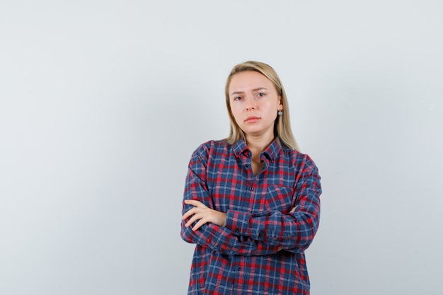 Mujer rubia de pie con los brazos cruzados en camisa a cuadros y mirando serio, vista frontal.