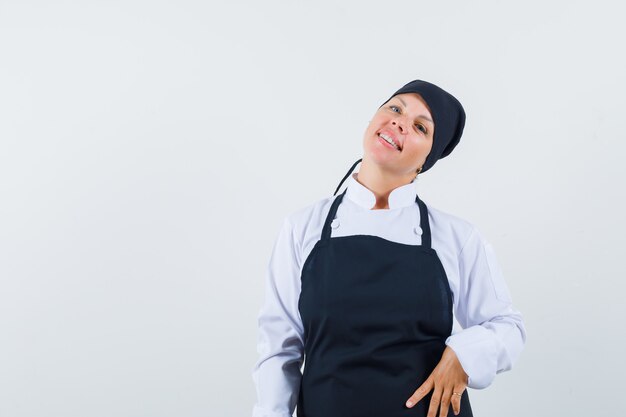 Mujer rubia de pie con la espalda recta, poniendo la mano en la cintura y posando al frente con uniforme de cocinero negro y muy bonita.