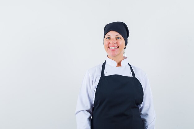 Mujer rubia de pie con la espalda recta y posando al frente con uniforme de cocinero negro y muy bonita.