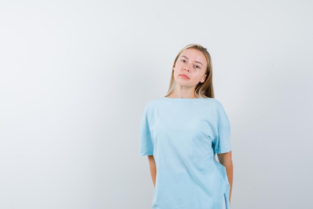 Mujer rubia de pie con la espalda recta y posando a la cámara en camiseta azul y mirando seria