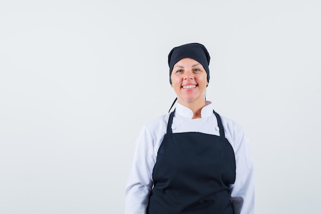 Mujer rubia de pie con la espalda recta, sonriendo y posando a la cámara en uniforme de cocinero negro y luciendo bonita. vista frontal.