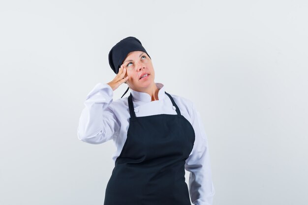 Mujer rubia de pie en pose de pensamiento, apoyando la mejilla en la mano con uniforme de cocinero negro y mirando pensativo.