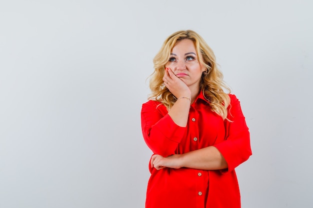 Mujer rubia de pie en pose de pensamiento en blusa roja y mirando pensativo.