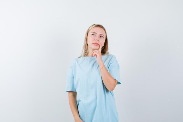 Mujer rubia de pie en pose de pensamiento, poniendo el dedo índice cerca de la boca en camiseta azul