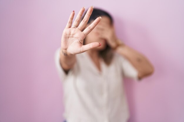 Mujer rubia de pie sobre fondo rosa cubriendo los ojos con las manos y haciendo un gesto de alto con expresión de tristeza y miedo. concepto avergonzado y negativo.