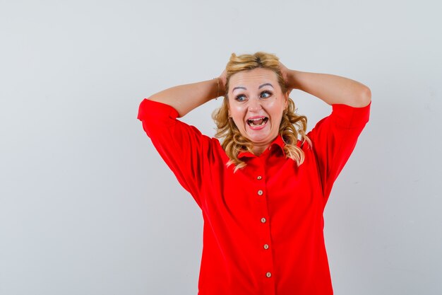 Mujer rubia poniendo las manos en la cabeza en blusa roja y mirando feliz.