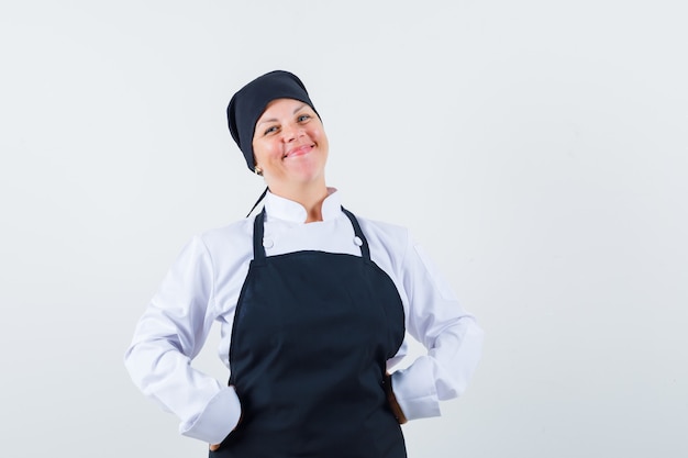 Mujer rubia poniendo las manos en la cintura en uniforme de cocinero negro y luciendo bonita vista frontal.