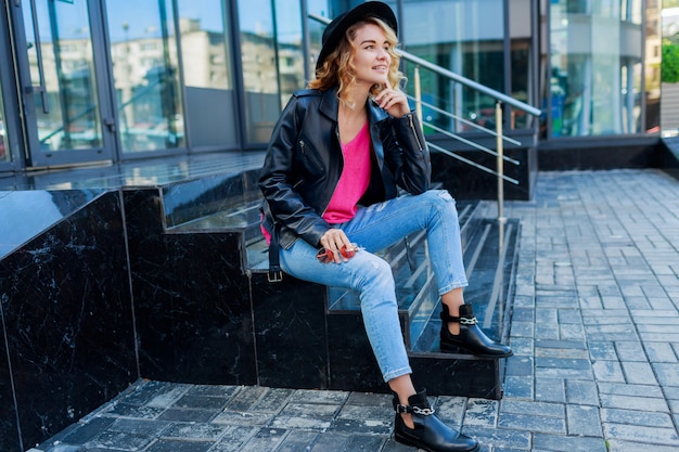 Mujer rubia posando en las calles modernas. Elegante traje de otoño, chaqueta de cuero y suéter de punto. Gafas de sol rosas