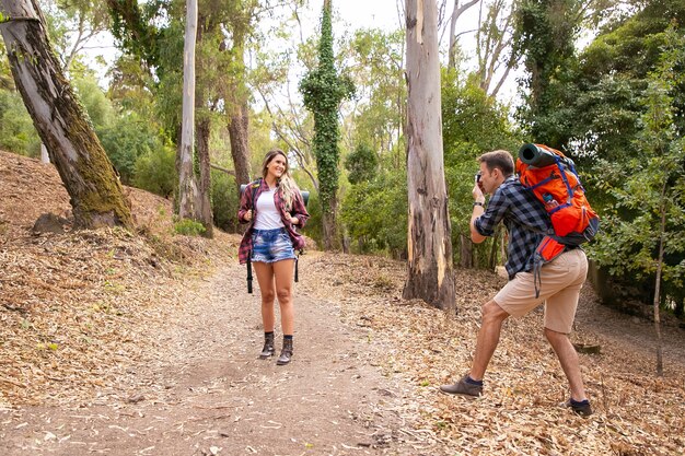 Mujer rubia posando para la foto en la carretera en el bosque. Hombre caucásico sosteniendo la cámara y disparando sobre la naturaleza. Dos personas felices haciendo senderismo con mochilas. Concepto de turismo, aventura y vacaciones de verano.