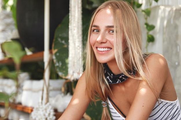 Mujer rubia positiva con amplia sonrisa, tiene una sonrisa brillante, está de buen humor mientras se recrea en la acogedora cafetería con su amante o amigo, vestida de manera informal, descansa después de caminar al aire libre. Felicidad