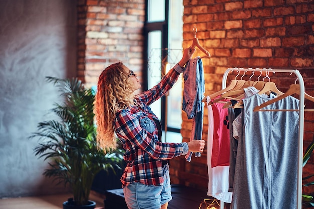 Mujer rubia positiva con anteojos negros elige ropa de moda en el perchero.