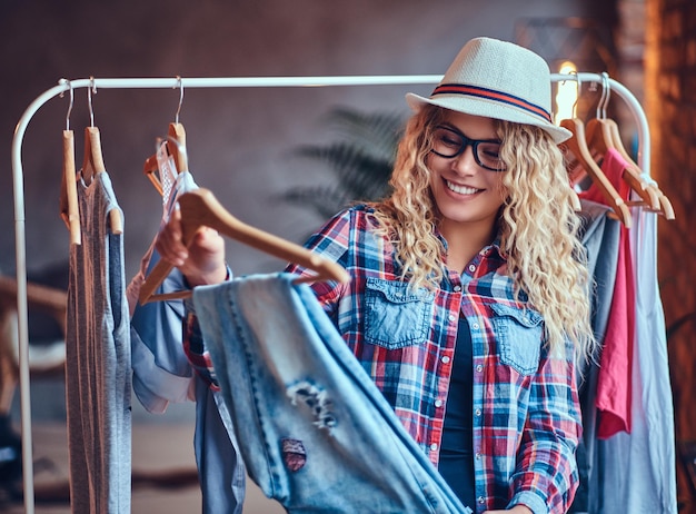 Mujer rubia positiva con anteojos negros elige ropa de moda en el perchero.