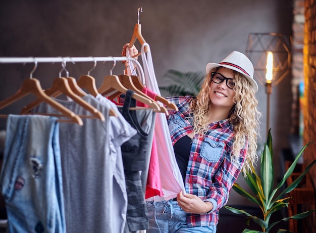 Mujer rubia positiva con anteojos negros elige ropa de moda en el perchero.