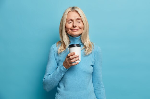 Mujer rubia positiva está parado con los ojos cerrados bebidas café para llevar