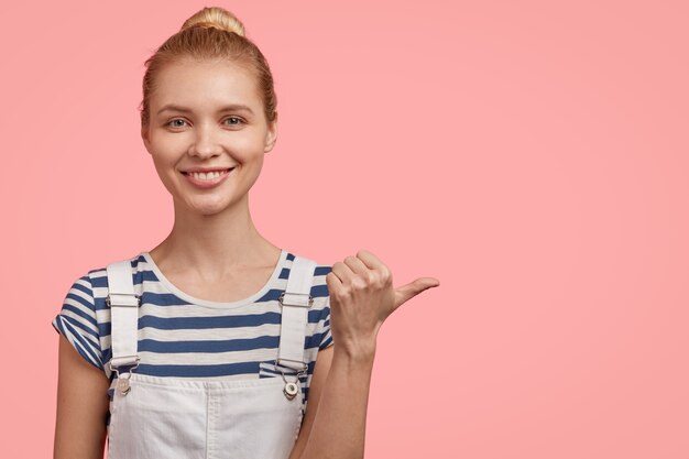 Mujer rubia positiva y satisfecha con el pelo peinado, viste una camiseta a rayas y un mono de mezclilla, señala con el pulgar a un lado, sonríe agradablemente, muestra el espacio libre derecho aislado sobre una pared rosa