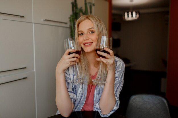 Mujer rubia prueba dos vasos de vino tinto en su propia cocina.