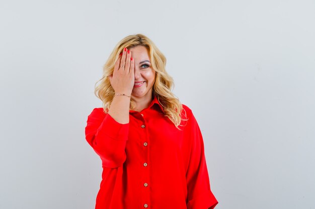 Mujer rubia que cubre los ojos con las manos en blusa roja y parece feliz,