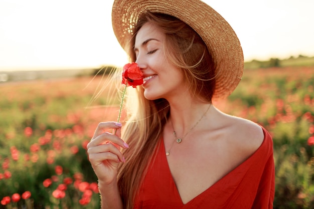 Mujer rubia romántica con flor en mano caminando en el increíble campo de amapolas. Colores cálidos del atardecer. Sombrero de copa. Vestido rojo. Colores suaves.
