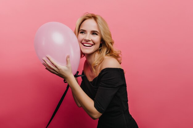 Mujer rubia romántica con gran sonrisa posando en vestido negro en la pared de color rosa brillante. encantadora cumpleañera con globo de helio.