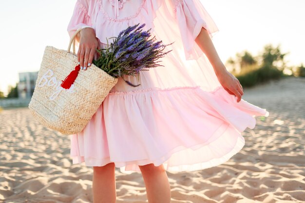 Mujer rubia romántica en lindo vestido rosa bailando y teniendo fu en la playa. Sosteniendo una bolsa de paja y un ramo de lavanda. Concepto de libertad y naturaleza.