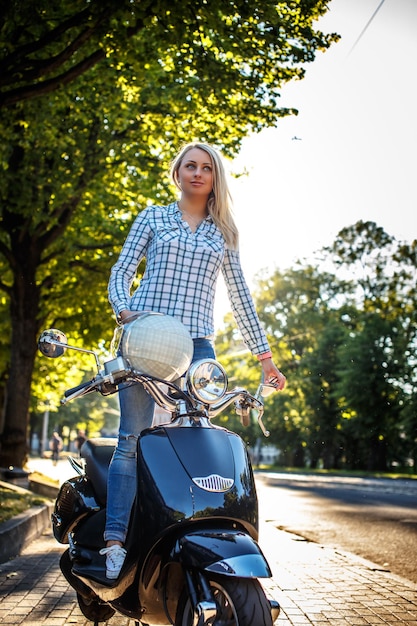 Mujer rubia con ropa informal posando en moto scooter sobre fondo de árboles verdes.