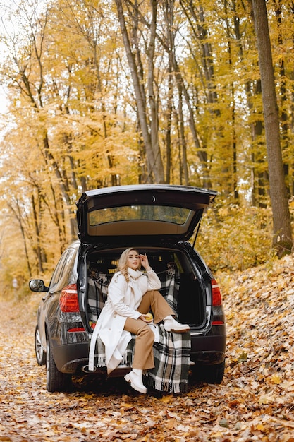 Mujer rubia sentada en el maletero de un coche negro en el bosque de otoño. Mujer con bata blanca. Chica sentada sobre una manta a cuadros.