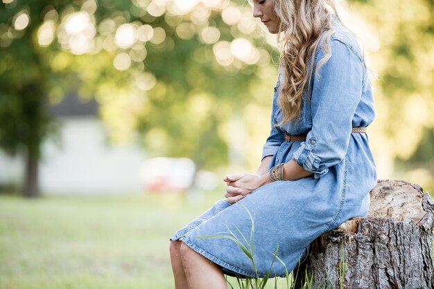 Mujer rubia sentada en el tocón de un árbol y rezando en un jardín bajo la luz del sol