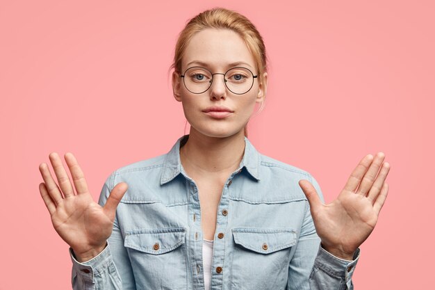 Mujer rubia seria con expresión segura, muestra las palmas de las manos, hace un gesto de parada, no permite hacer algo, usa ropa de moda, aislada en la pared rosa