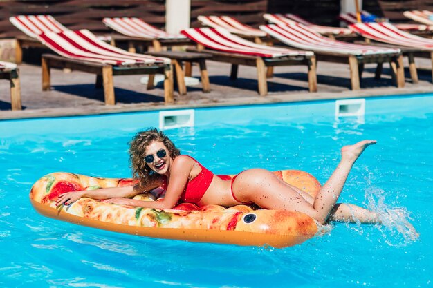 Mujer rubia sexual con culo en forma acostado sobre pizza flotante en la piscina. Tiempo feliz, vacaciones de verano.