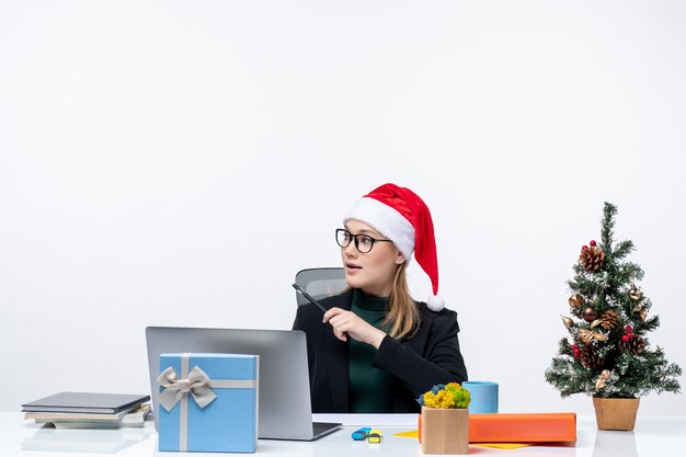 Mujer rubia con un sombrero de santa claus sentado en una mesa con un árbol de Navidad y un regalo