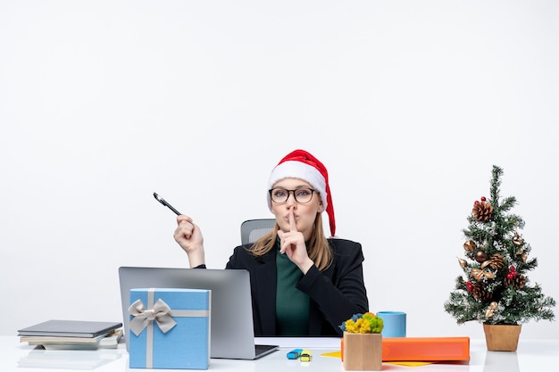 Mujer rubia con un sombrero de santa claus sentado en una mesa con un árbol de Navidad y un regalo