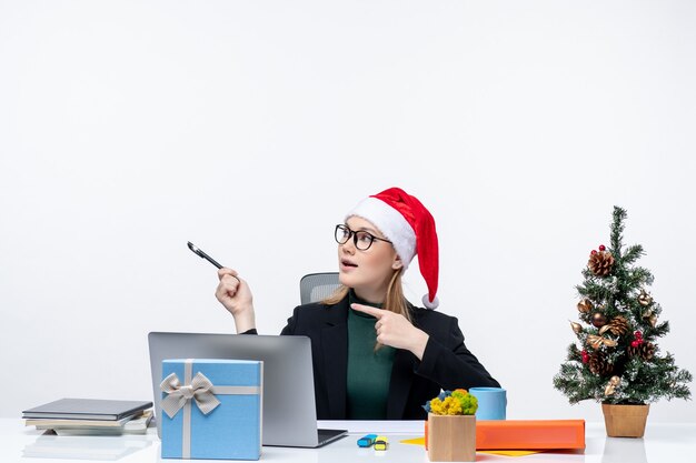 Mujer rubia con un sombrero de santa claus sentado en una mesa con un árbol de Navidad y un regalo