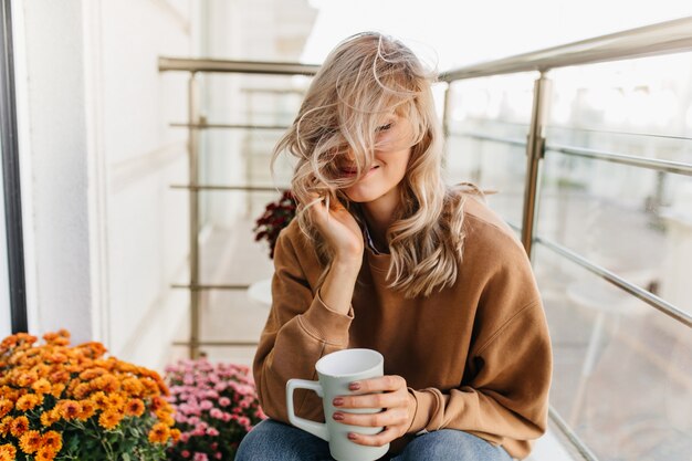 Mujer rubia soñadora sentada en el balcón con una taza de té. Impresionante modelo femenina relajándose en la terraza.