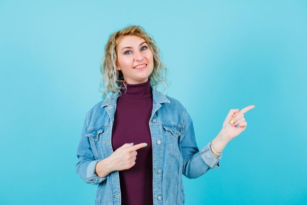 Mujer rubia sonriente apunta a la derecha con los dedos índices sobre fondo azul.