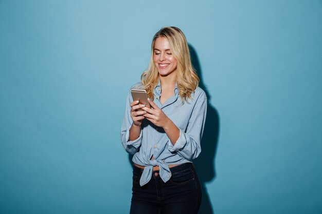 Mujer rubia sonriente en camisa escribiendo mensaje en teléfono inteligente