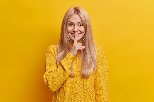 Mujer rubia sonriente complacida hace gesto secreto, dedo en el labio, callando