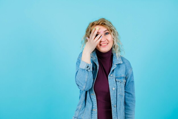 Mujer rubia sonriente está sosteniendo los dedos en el ojo sobre fondo azul.