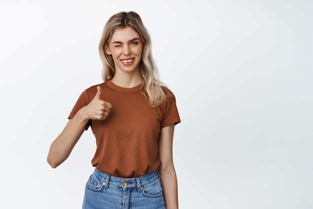 Mujer rubia sonriente guiñando un ojo mostrando el pulgar hacia arriba en señal de aprobación como algo de pie en camiseta marrón y jeans sobre fondo blanco.