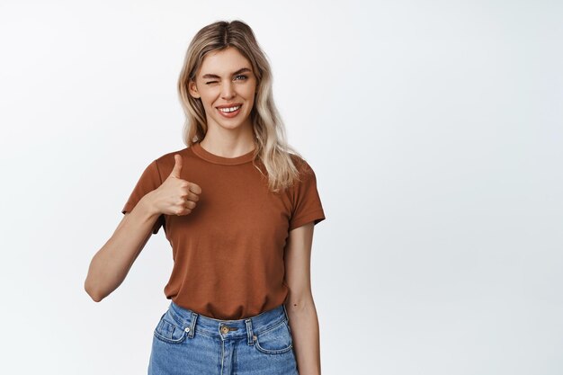 Mujer rubia sonriente guiñando un ojo mostrando el pulgar hacia arriba en señal de aprobación como algo de pie en camiseta marrón y jeans sobre fondo blanco.