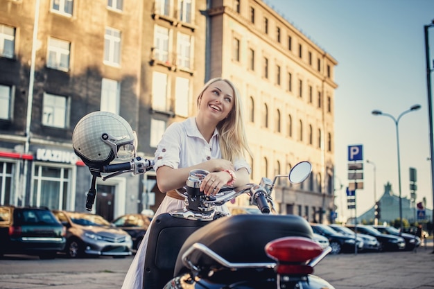 Mujer rubia sonriente con vestido blanco posando cerca de moto scooter.