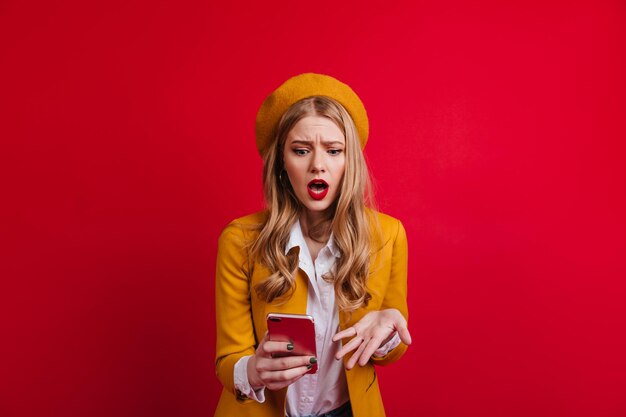 Mujer rubia sorprendida en boina mirando el teléfono inteligente Chica francesa molesta de pie sobre fondo rojo con la boca abierta