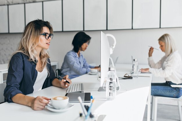 Mujer rubia sorprendida mirando la pantalla de la computadora y disfrutando del café con leche en su lugar de trabajo