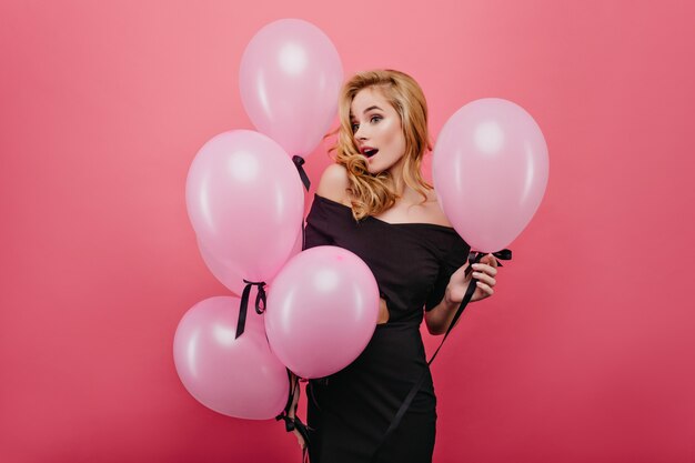 Mujer rubia sorprendida sosteniendo globos de fiesta hermosos. asombrada chica adorable en traje negro aislado en la pared rosa.